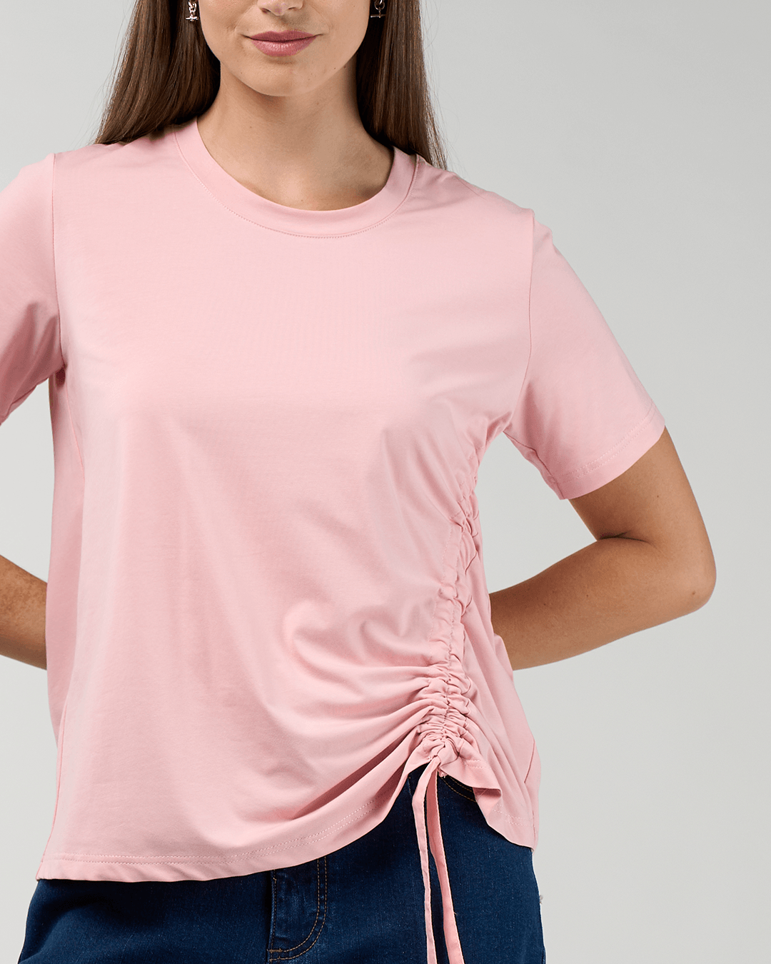 Madly Sweetly modeling a light pink short-sleeve t-shirt with a side ruching detail and tie, paired with dark blue jeans, against a light gray studio background.