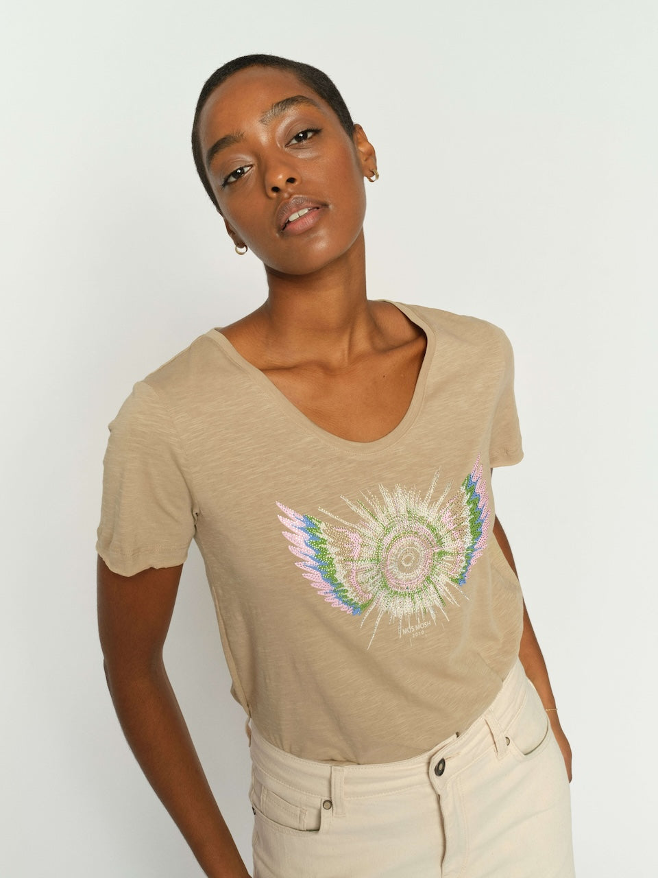 Mos Mosh  woman modeling a beige short-sleeve t-shirt with a colorful embroidered angel wing design on the front, paired with cream-colored pants, against a plain light gray background.
