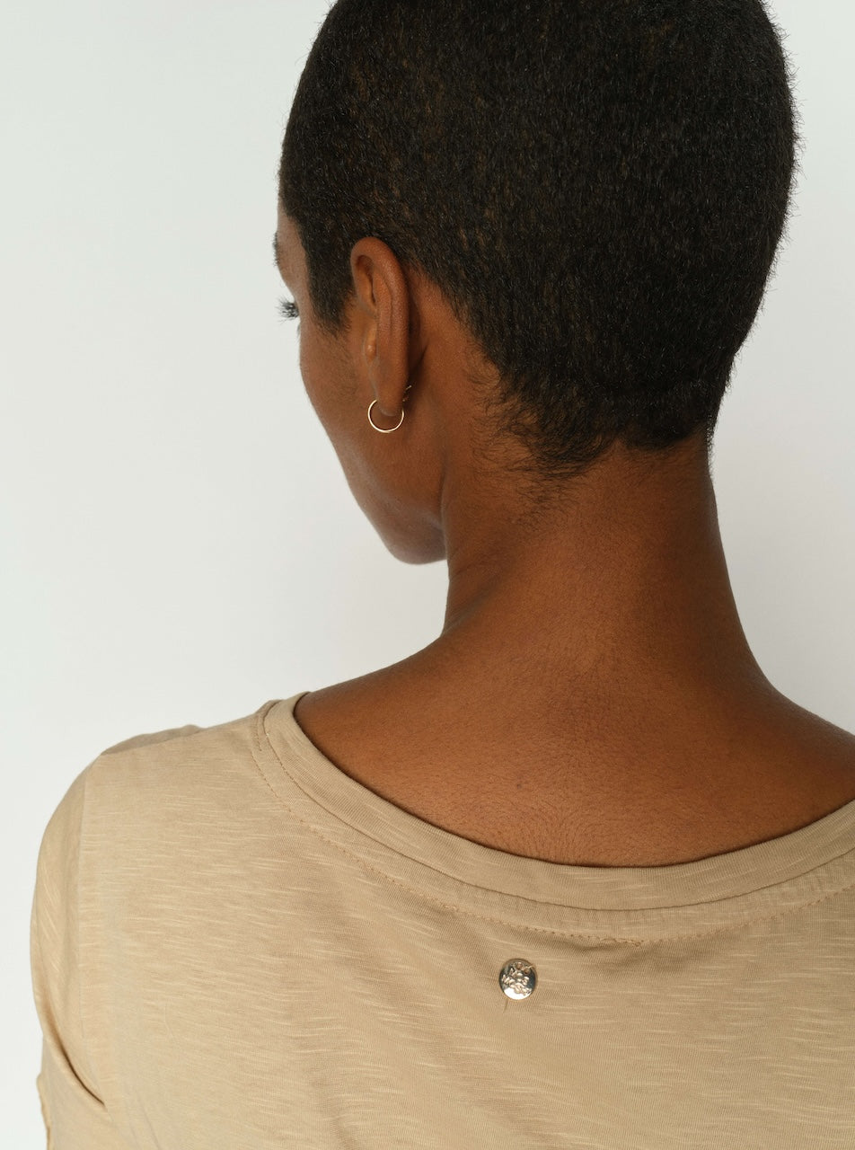 A close-up back view of a person with short dark hair wearing a beige crew-neck top with a small silver button detail at the back neckline, against a plain light background.