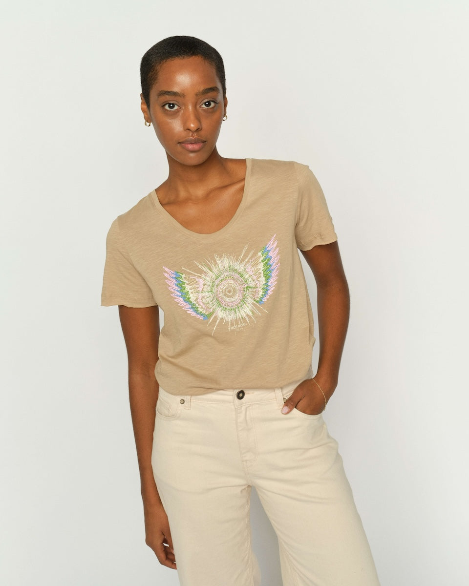 Mos Mosh  woman modeling a beige short-sleeve t-shirt with a colorful embroidered angel wing design on the front, paired with cream-colored pants, against a plain light gray background.