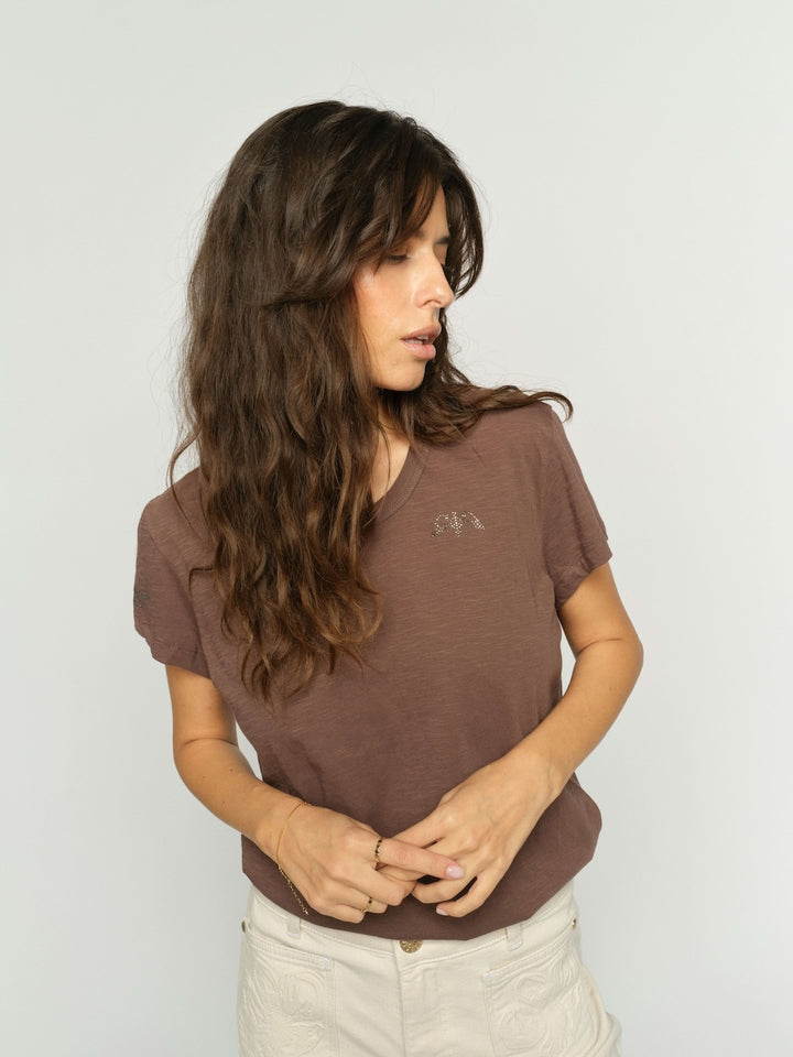 Mos Mosh woman with long, wavy brown hair wearing a brown short-sleeve V-neck t-shirt with a small embroidered logo on the left chest, paired with light beige pants, photographed against a light gray studio background.