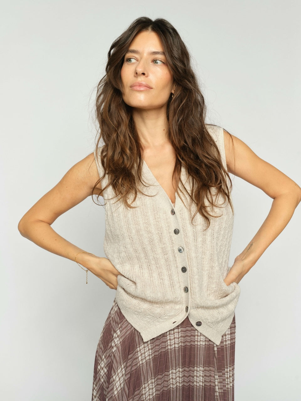 Mos Mosh  woman modeling a sleeveless, cable-knit vest in a light beige color with a button-down front, paired with a brown plaid skirt, against a plain light gray background.