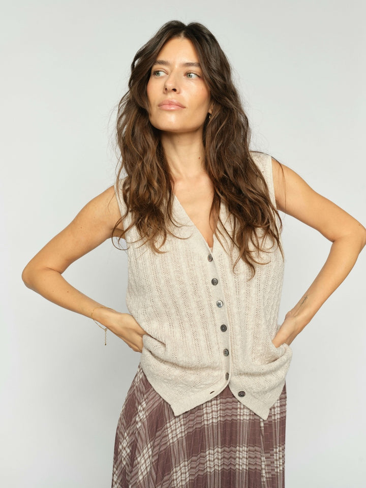 Mos Mosh  woman modeling a sleeveless, cable-knit vest in a light beige color with a button-down front, paired with a brown plaid skirt, against a plain light gray background.