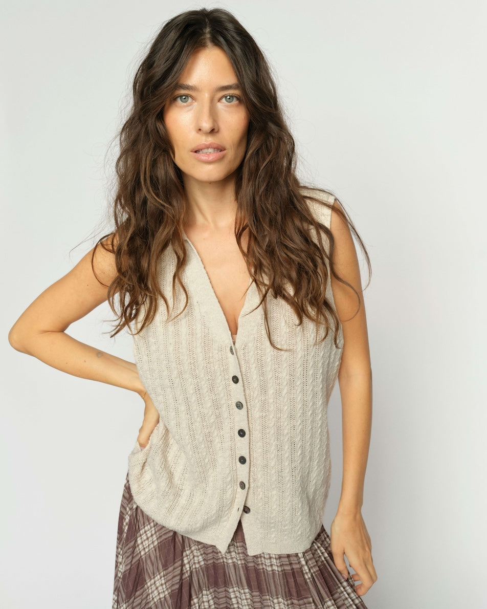 Mos Mosh  woman modeling a sleeveless, cable-knit vest in a light beige color with a button-down front, paired with a brown plaid skirt, against a plain light gray background.
