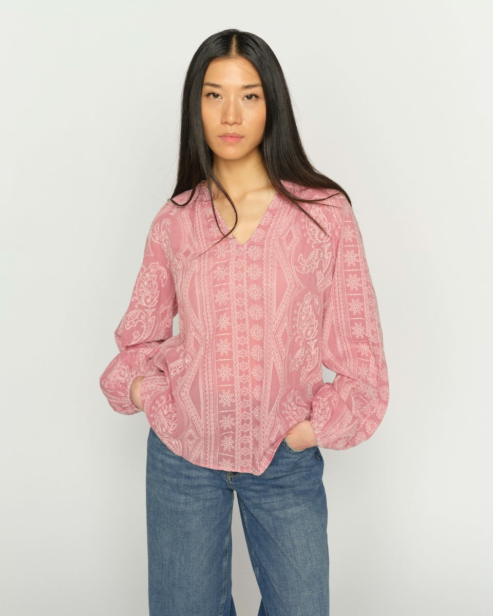 A woman modeling a pink patterned blouse with blue jeans, standing against a plain light gray background.