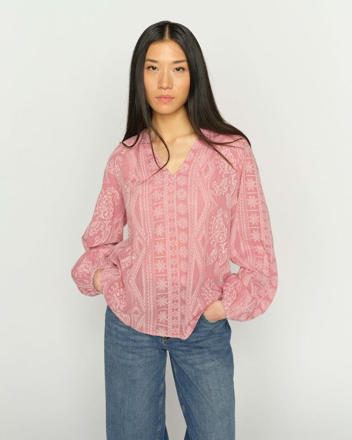 A woman modeling a pink patterned blouse with blue jeans, standing against a plain light gray background.