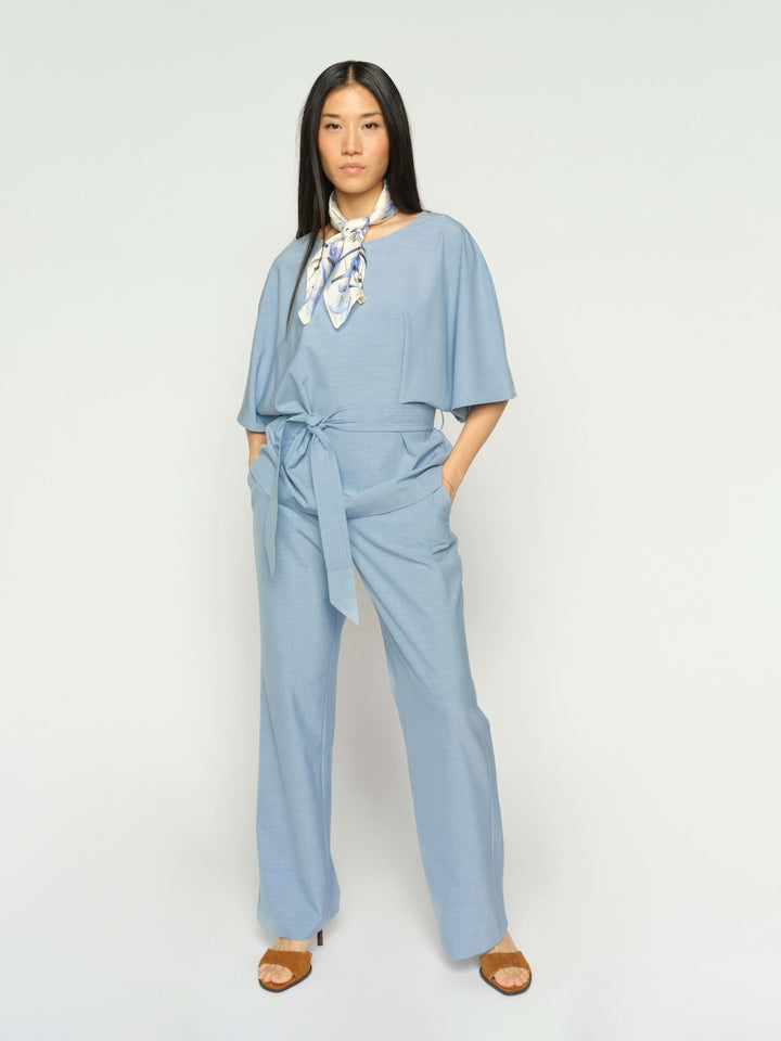Mos Mosh woman modeling a white long-sleeve blouse with metallic gold leaf embroidery on the shoulders, paired with light blue wide-leg trousers and beige sandals, against a solid light gray studio background.