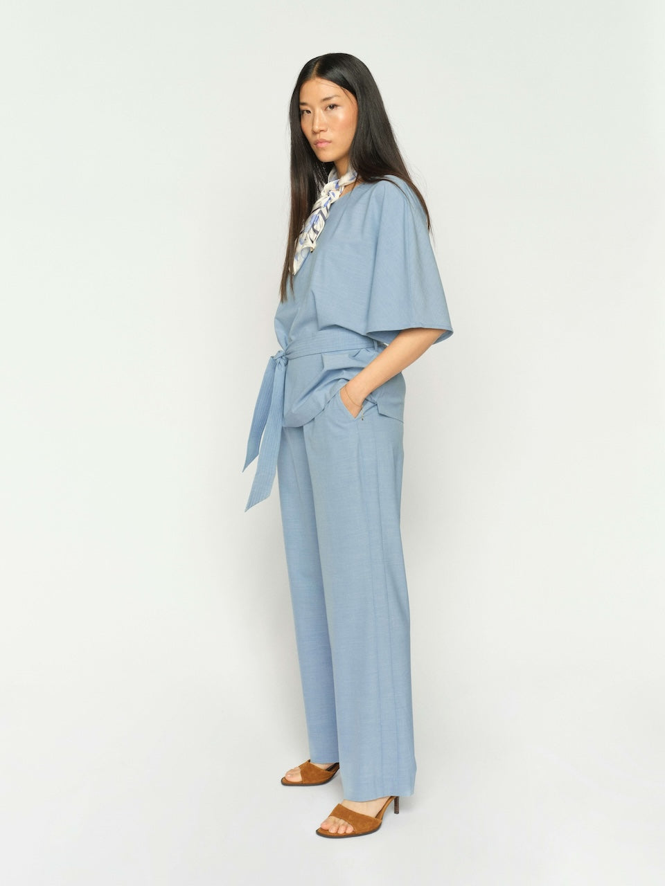 Mos Mosh woman modeling a white long-sleeve blouse with metallic gold leaf embroidery on the shoulders, paired with light blue wide-leg trousers and beige sandals, against a solid light gray studio background.