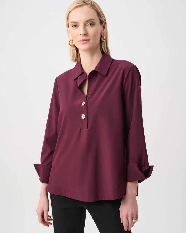 Joseph Ribkoff  modeling a long-sleeve button-front blouse in deep burgundy with a collar and rolled cuffs, paired with black pants, against a plain light gray studio background.