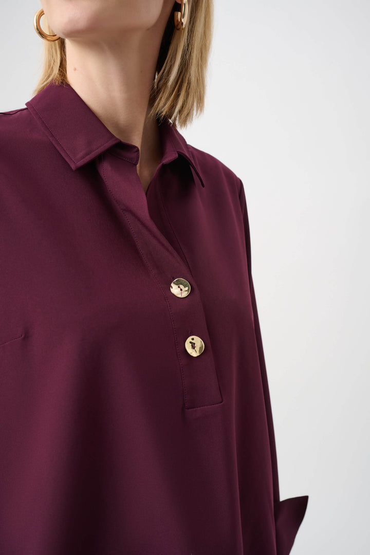 Joseph Ribkoff  modeling a long-sleeve button-front blouse in deep burgundy with a collar and rolled cuffs, paired with black pants, against a plain light gray studio background. Close up