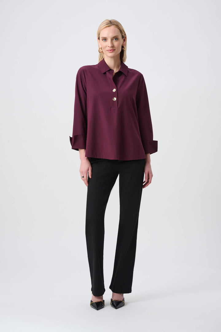 Joseph Ribkoff  modeling a long-sleeve button-front blouse in deep burgundy with a collar and rolled cuffs, paired with black pants, against a plain light gray studio background.