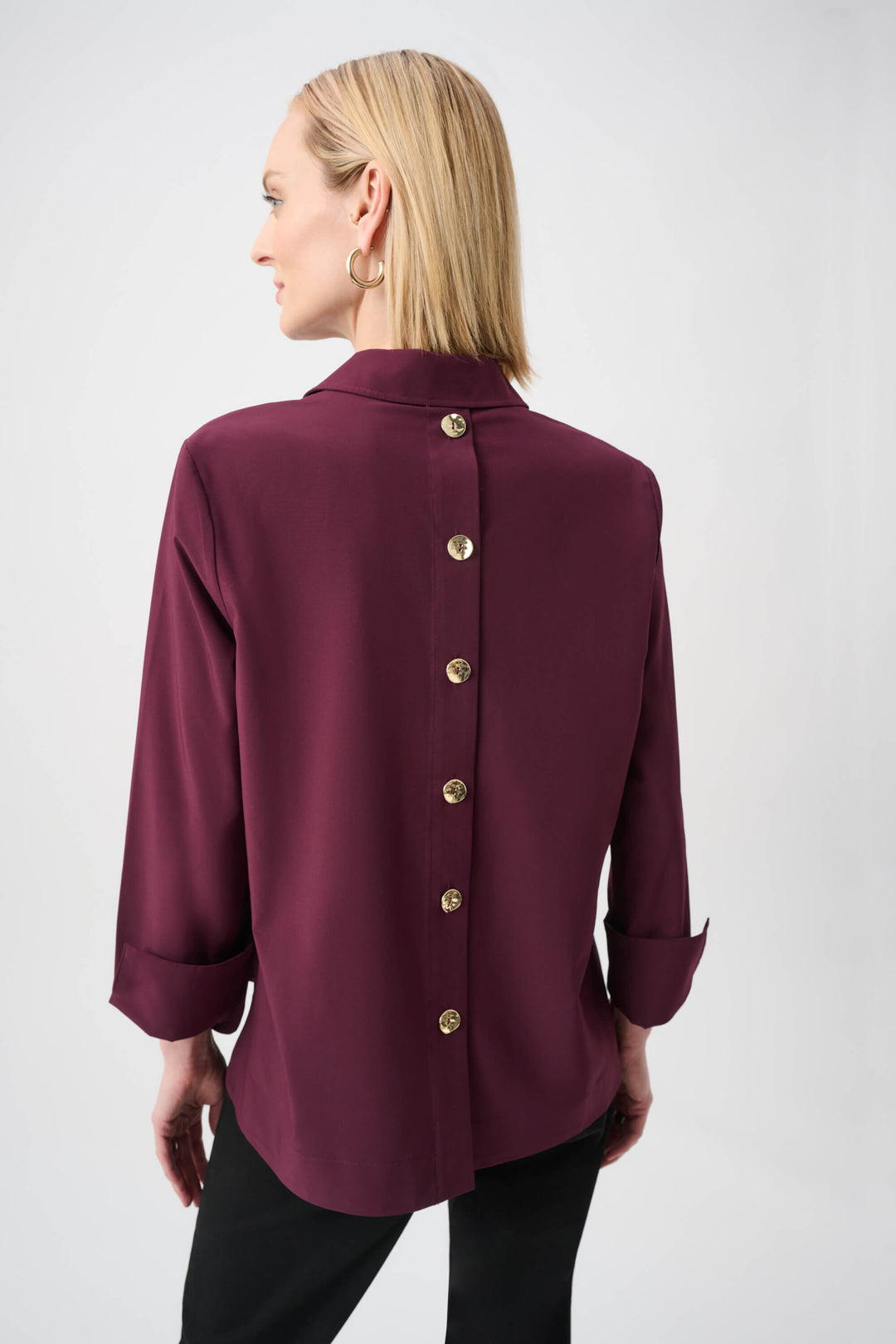 Joseph Ribkoff  modeling a long-sleeve button-front blouse in deep burgundy with a collar and rolled cuffs, paired with black pants, against a plain light gray studio background. Back view