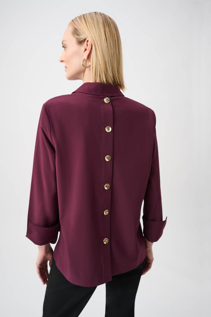 Joseph Ribkoff  modeling a long-sleeve button-front blouse in deep burgundy with a collar and rolled cuffs, paired with black pants, against a plain light gray studio background. Back view
