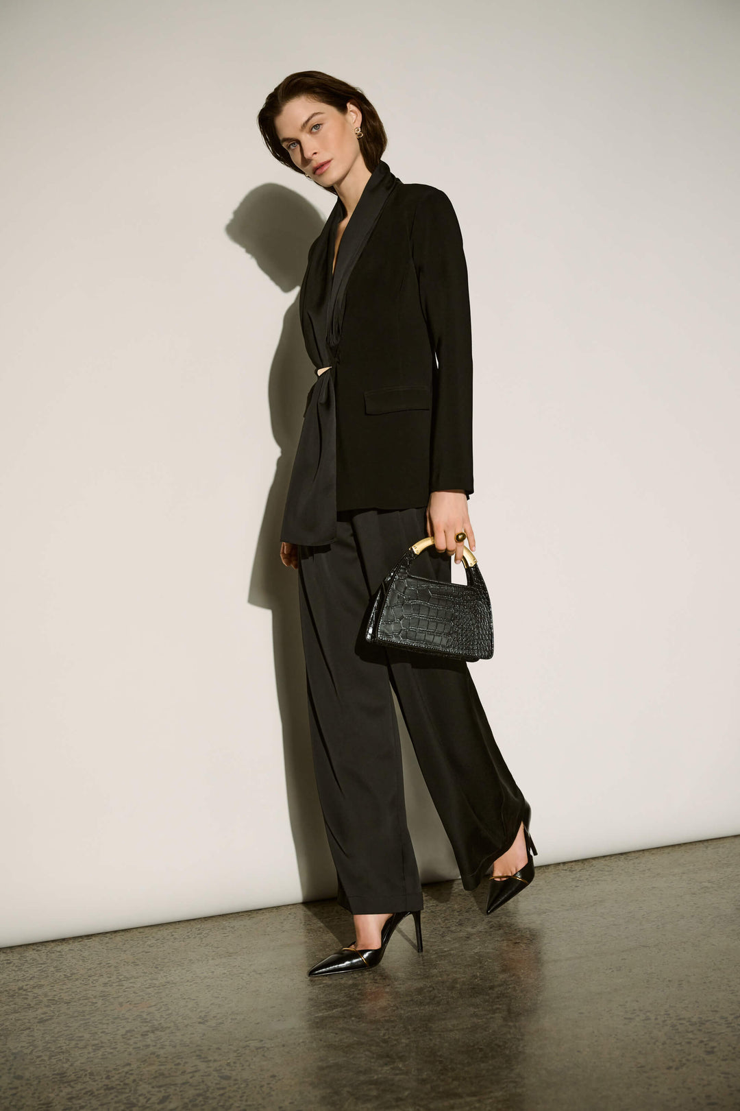 Joseph Ribkoff  model wearing a black tailored blazer and matching wide-leg trousers, holding a small black textured handbag with gold-toned handles, posed against a minimalist off-white wall with dramatic shadows.