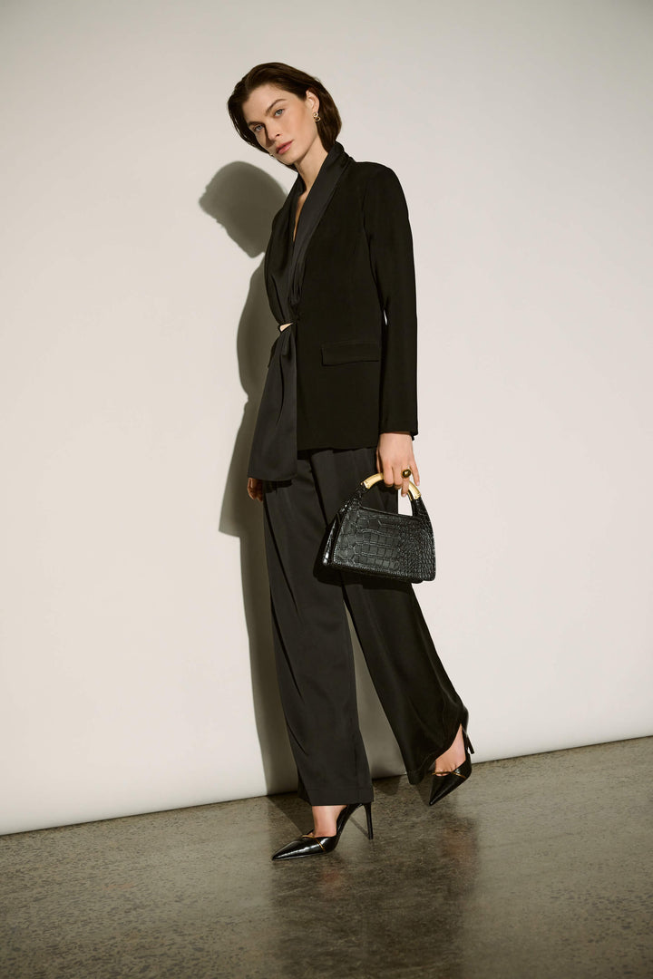 Joseph Ribkoff  model wearing a black tailored blazer and matching wide-leg trousers, holding a small black textured handbag with gold-toned handles, posed against a minimalist off-white wall with dramatic shadows.