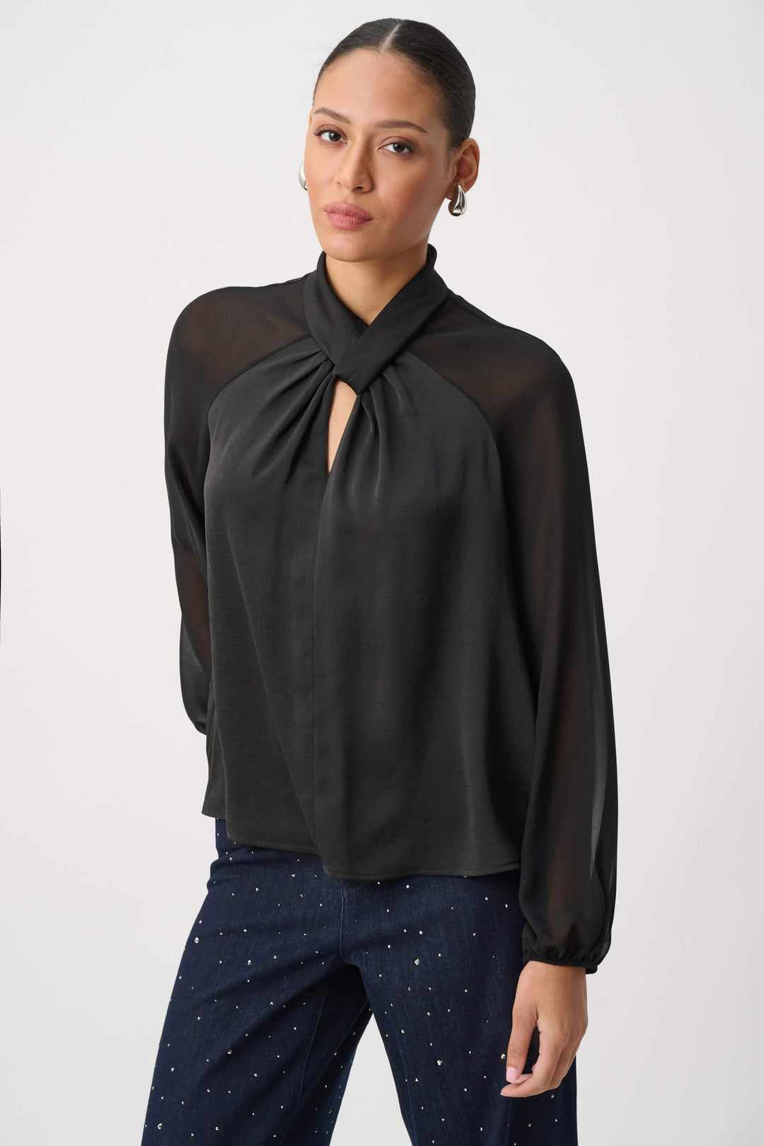 Joseph Ribkoff  woman wearing a black long-sleeve blouse with sheer mesh sleeves and a gathered keyhole-style neckline, paired with dark patterned pants, against a plain light gray studio background.