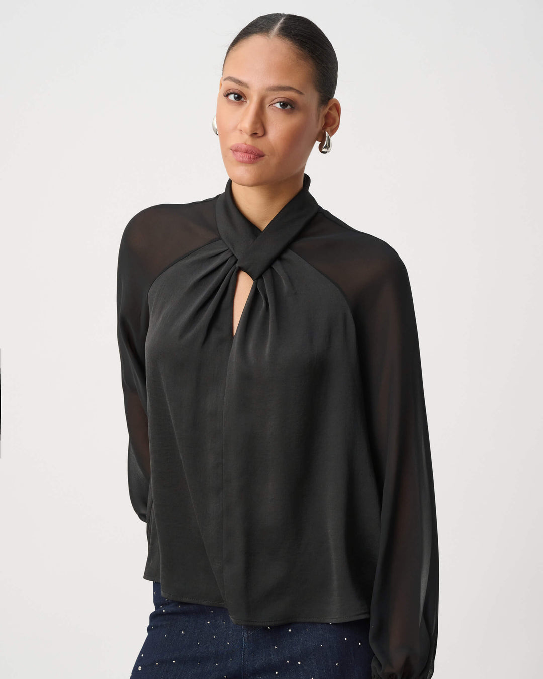 Joseph Ribkoff  woman wearing a black long-sleeve blouse with sheer mesh sleeves and a gathered keyhole-style neckline, paired with dark patterned pants, against a plain light gray studio background.