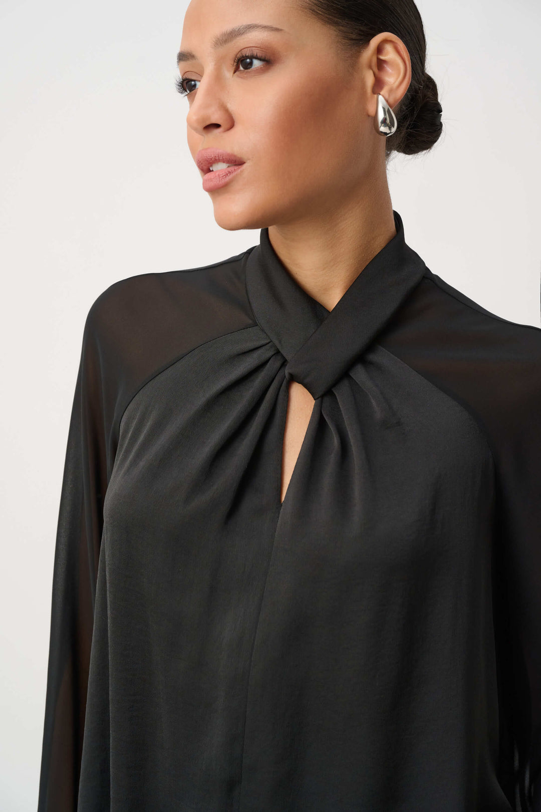 Joseph Ribkoff  woman wearing a black long-sleeve blouse with sheer mesh sleeves and a gathered keyhole-style neckline, paired with dark patterned pants, against a plain light gray studio background.