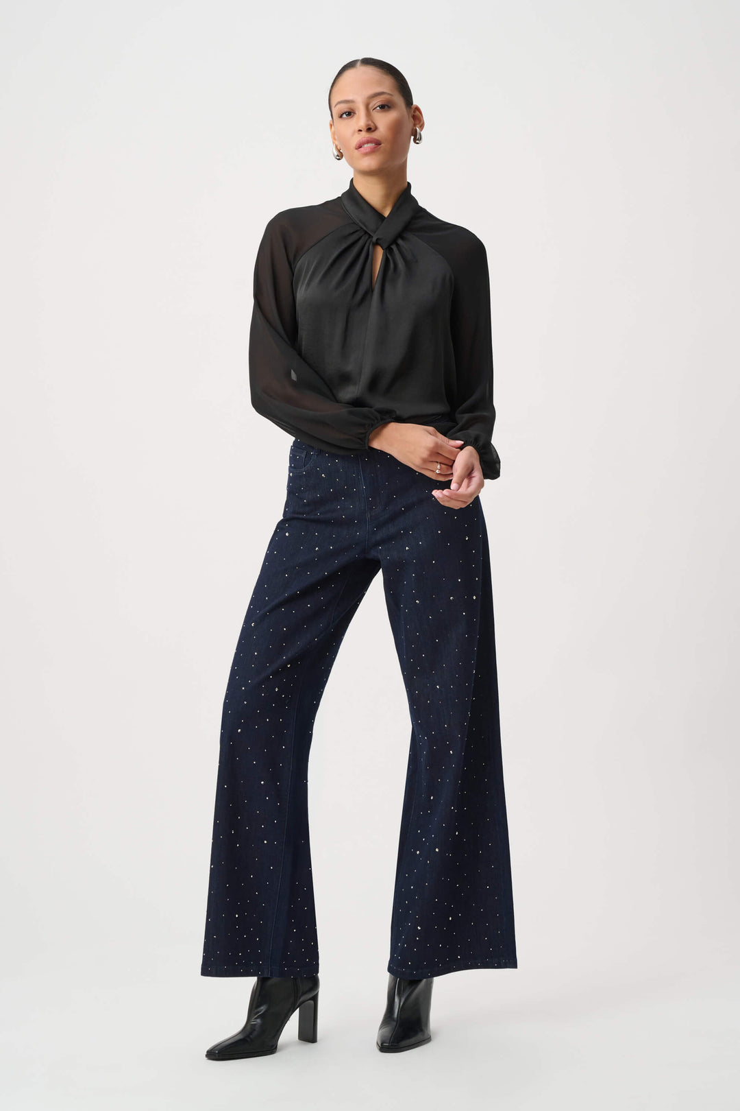 Joseph Ribkoff  woman wearing a black long-sleeve blouse with sheer mesh sleeves and a gathered keyhole-style neckline, paired with dark patterned pants, against a plain light gray studio background.