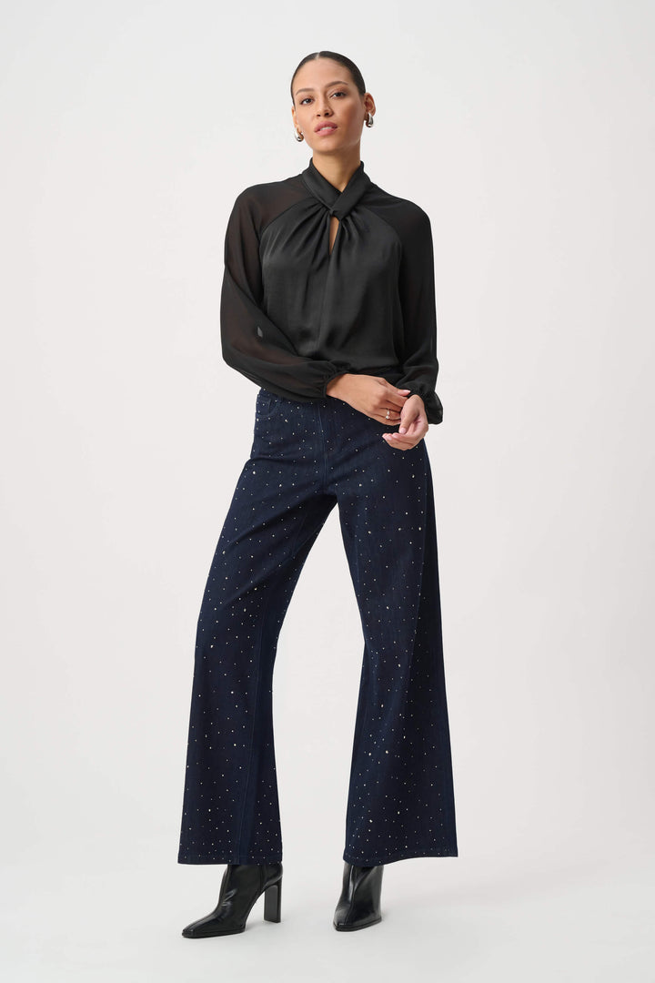 Joseph Ribkoff  woman wearing a black long-sleeve blouse with sheer mesh sleeves and a gathered keyhole-style neckline, paired with dark patterned pants, against a plain light gray studio background.
