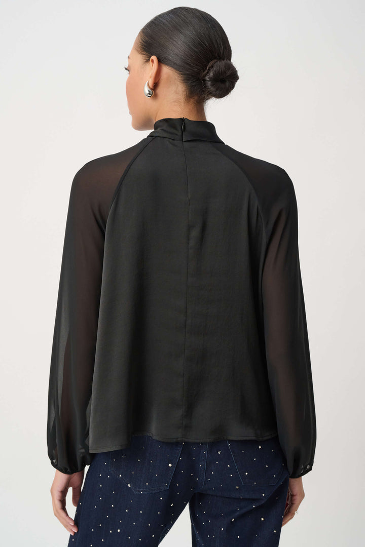 Joseph Ribkoff  woman wearing a black long-sleeve blouse with sheer mesh sleeves and a gathered keyhole-style neckline, paired with dark patterned pants, against a plain light gray studio background. Back view