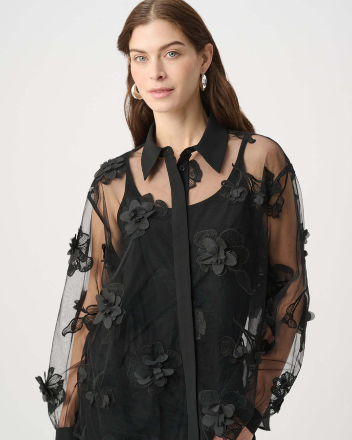 Joseph Ribkoff modeling a black sheer mesh blouse with 3D floral appliqués and a tie-neck collar, paired with black pants against a light gray studio background.
