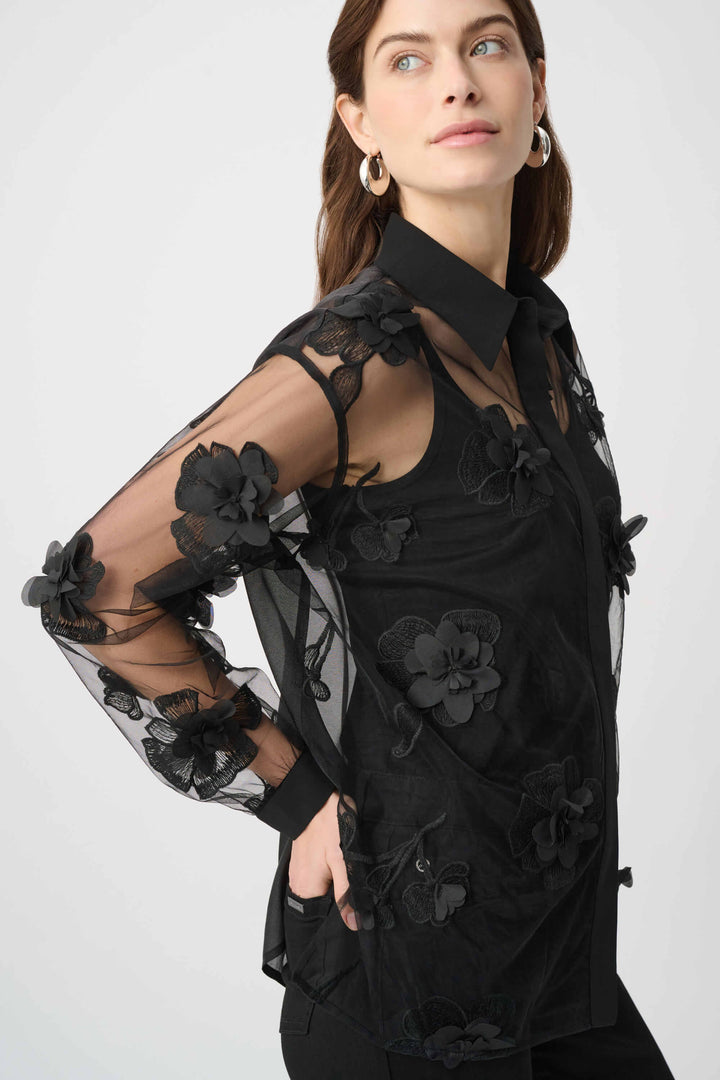 Joseph Ribkoff modeling a black sheer mesh blouse with 3D floral appliqués and a tie-neck collar, paired with black pants against a light gray studio background.