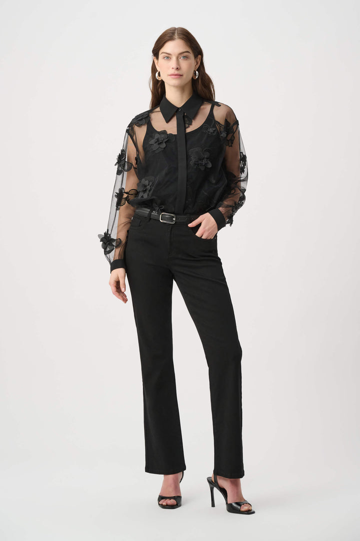 Joseph Ribkoff modeling a black sheer mesh blouse with 3D floral appliqués and a tie-neck collar, paired with black pants against a light gray studio background.