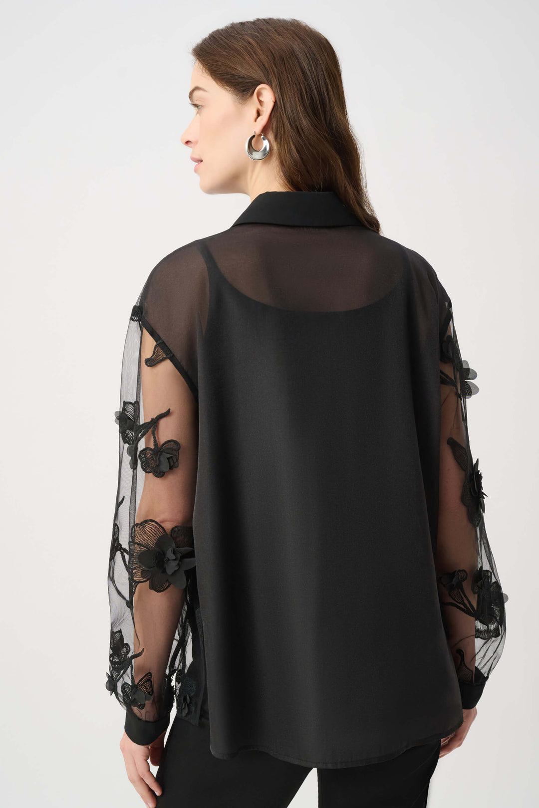 Joseph Ribkoff modeling a black sheer mesh blouse with 3D floral appliqués and a tie-neck collar, paired with black pants against a light gray studio background. Back view