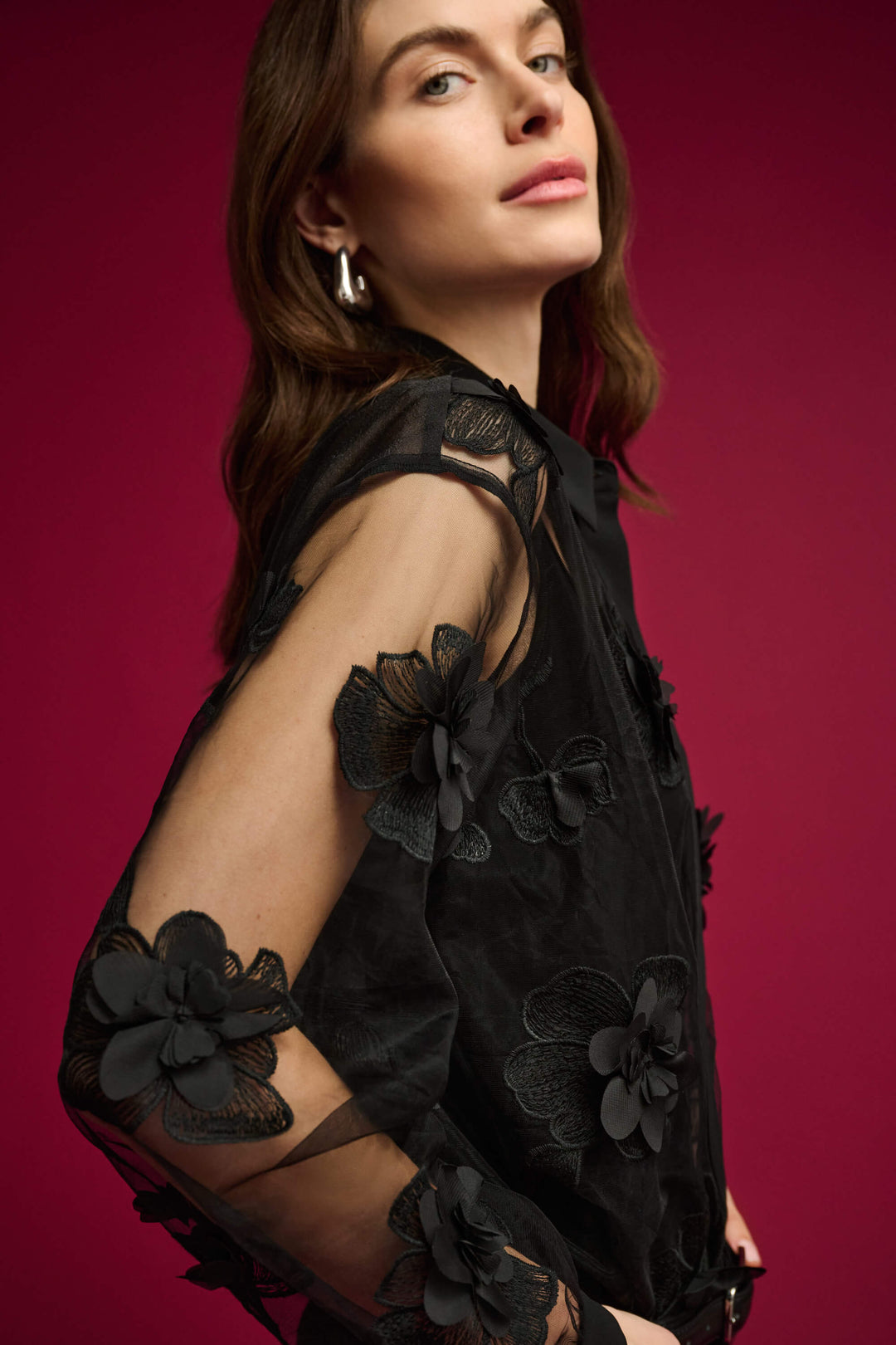 Joseph Ribkoff modeling a black sheer mesh blouse with 3D floral appliqués and a tie-neck collar, paired with black pants against a light gray studio background.