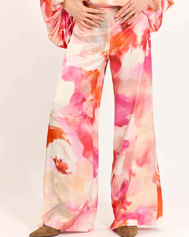 A person wearing wide-leg satin pants with a pink, orange, and white watercolor floral print, paired with brown pointed-toe flats, against a plain white studio background.