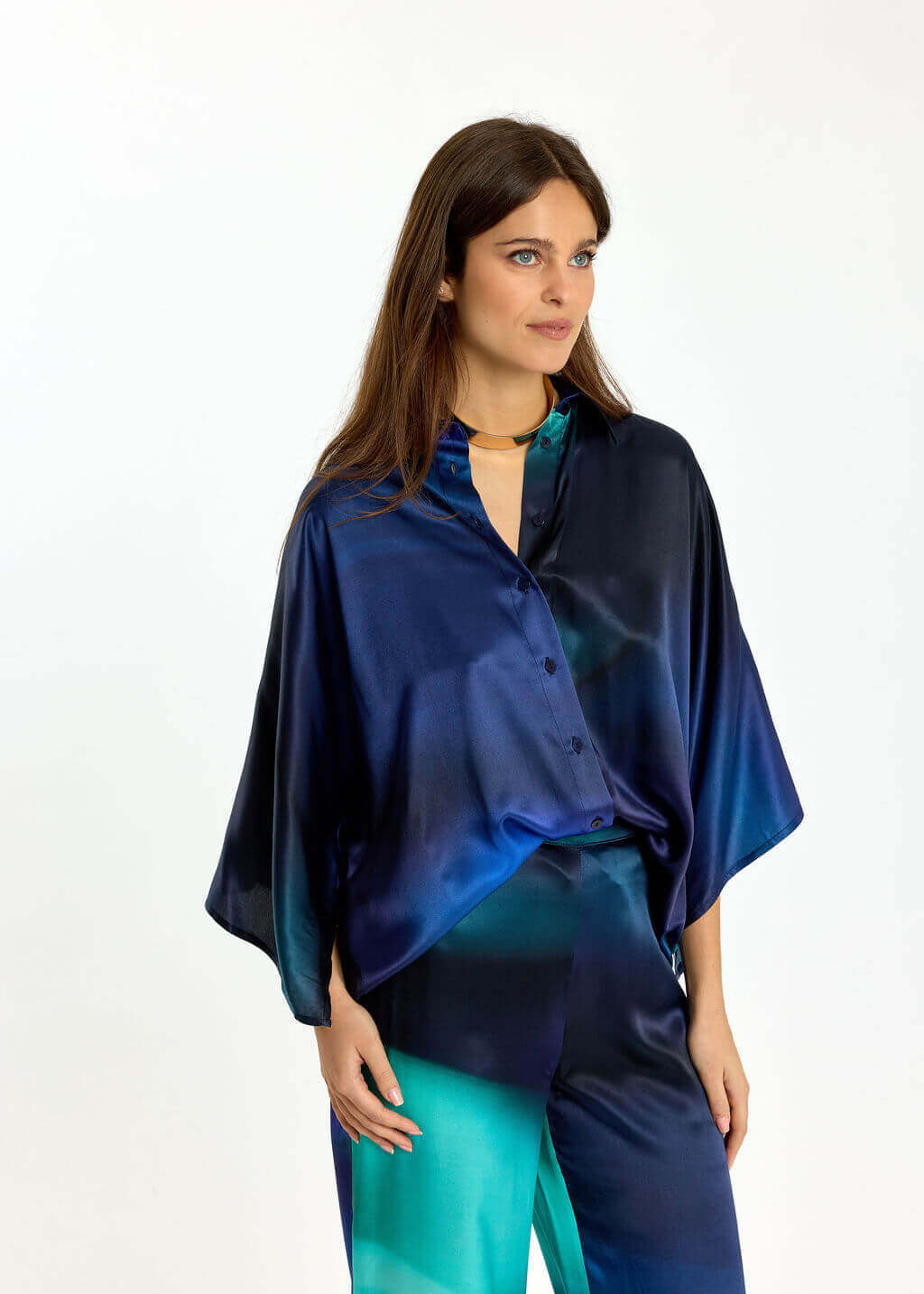Lauren Vidal  modeling a matching blue and teal satin button-up shirt and trousers set against a plain white studio background.