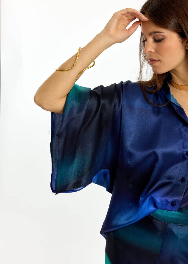 Lauren Vidal  modeling a matching blue and teal satin button-up shirt and trousers set against a plain white studio background.