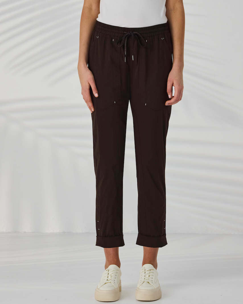 Lania The Label A model from the waist down wearing dark brown drawstring cropped pants with small metallic embellishments, paired with a white top and off-white sneakers, set against a white background with soft leaf shadows.
