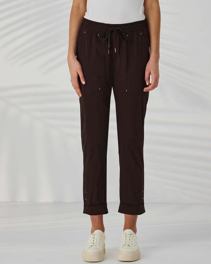 Lania The Label A model from the waist down wearing dark brown drawstring cropped pants with small metallic embellishments, paired with a white top and off-white sneakers, set against a white background with soft leaf shadows.