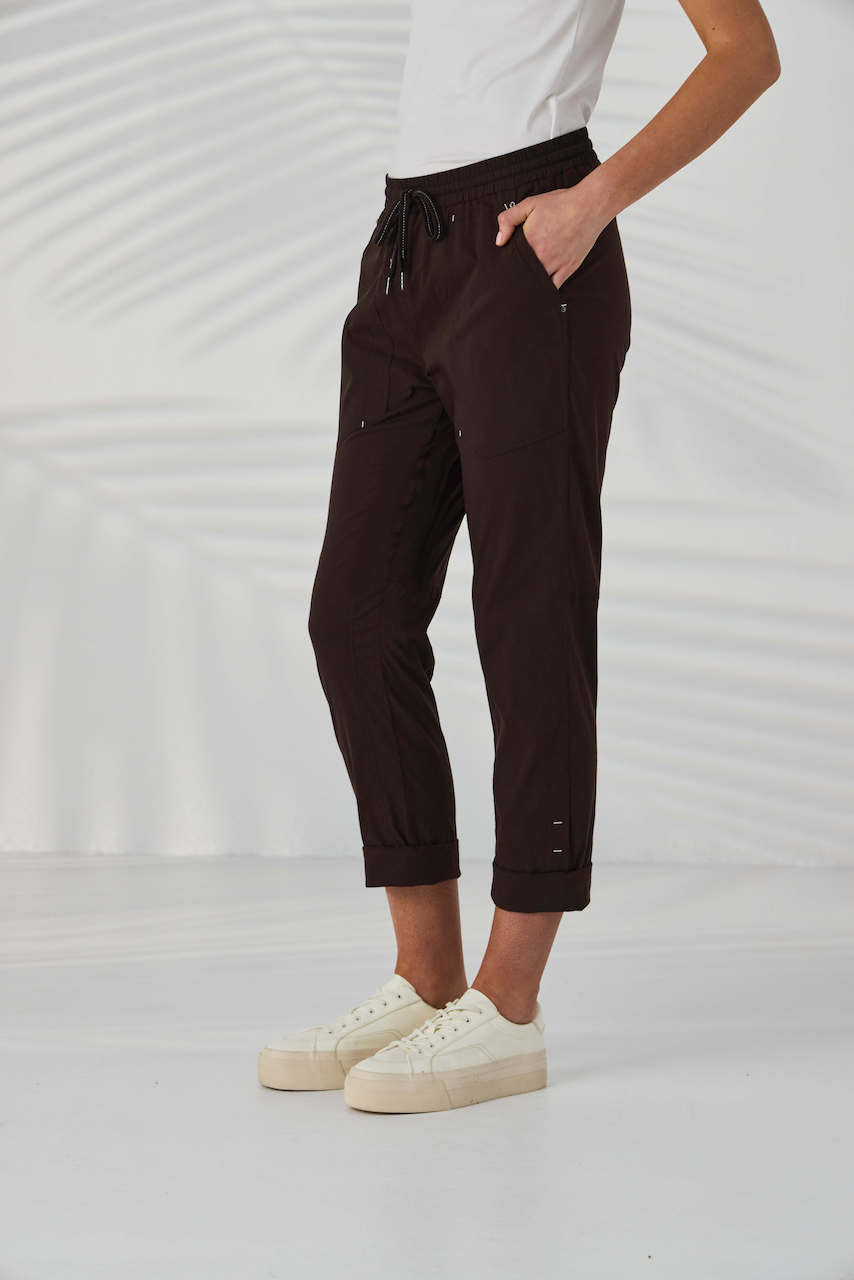 Lania The Label A model from the waist down wearing dark brown drawstring cropped pants with small metallic embellishments, paired with a white top and off-white sneakers, set against a white background with soft leaf shadows.