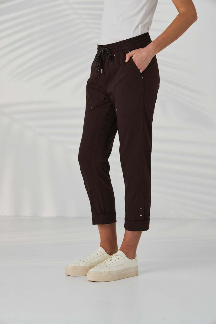 Lania The Label A model from the waist down wearing dark brown drawstring cropped pants with small metallic embellishments, paired with a white top and off-white sneakers, set against a white background with soft leaf shadows.