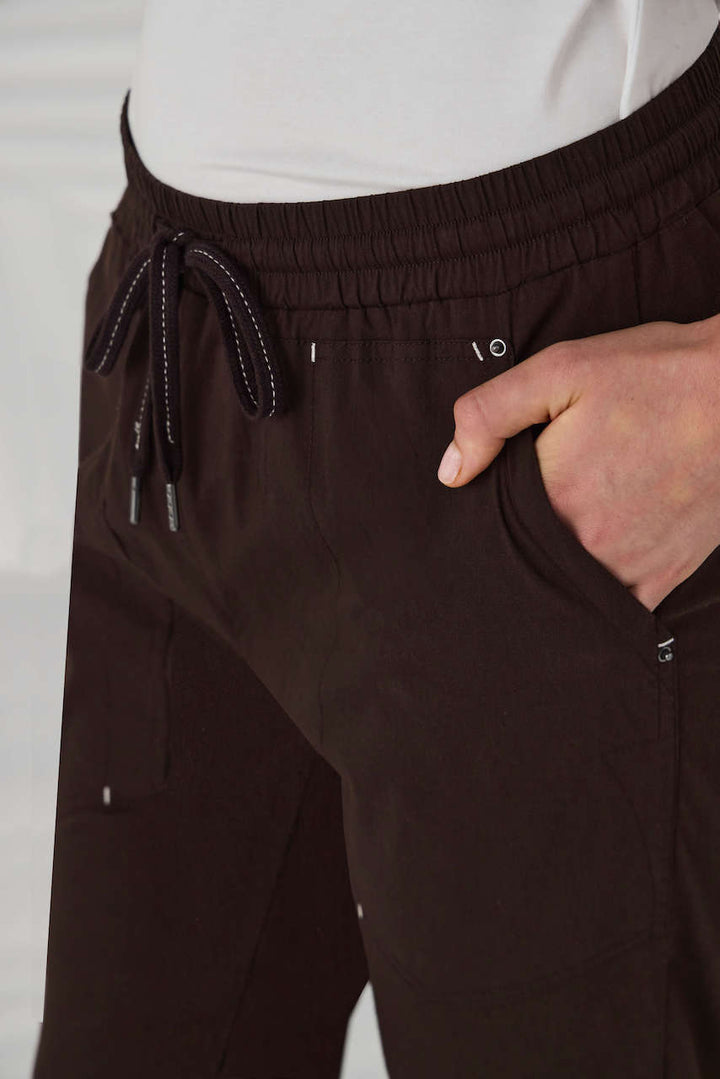 Lania The Label A model from the waist down wearing dark brown drawstring cropped pants with small metallic embellishments, paired with a white top and off-white sneakers, set against a white background with soft leaf shadows.