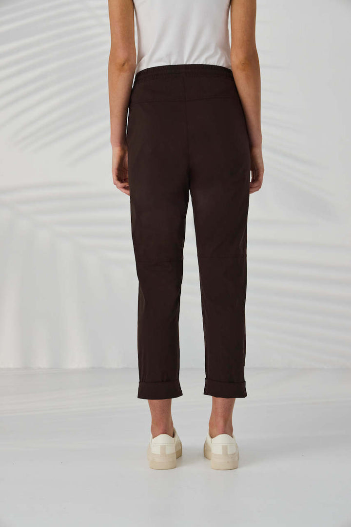 Lania The Label A model from the waist down wearing dark brown drawstring cropped pants with small metallic embellishments, paired with a white top and off-white sneakers, set against a white background with soft leaf shadows.