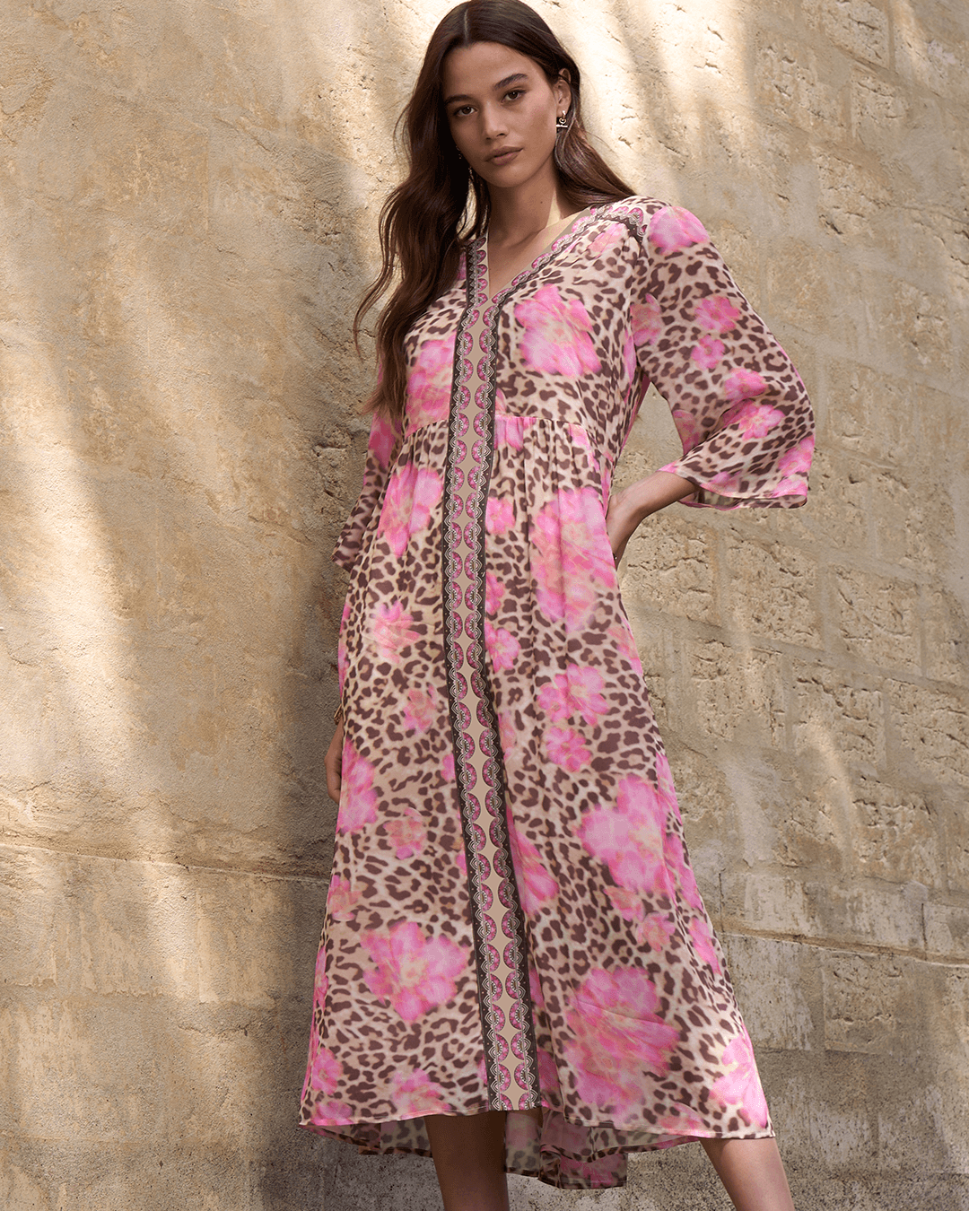 Loobie's Story models a long-sleeved midi dress with a pink and brown leopard print pattern overlaid with large pink floral motifs, standing against a textured beige stone wall.