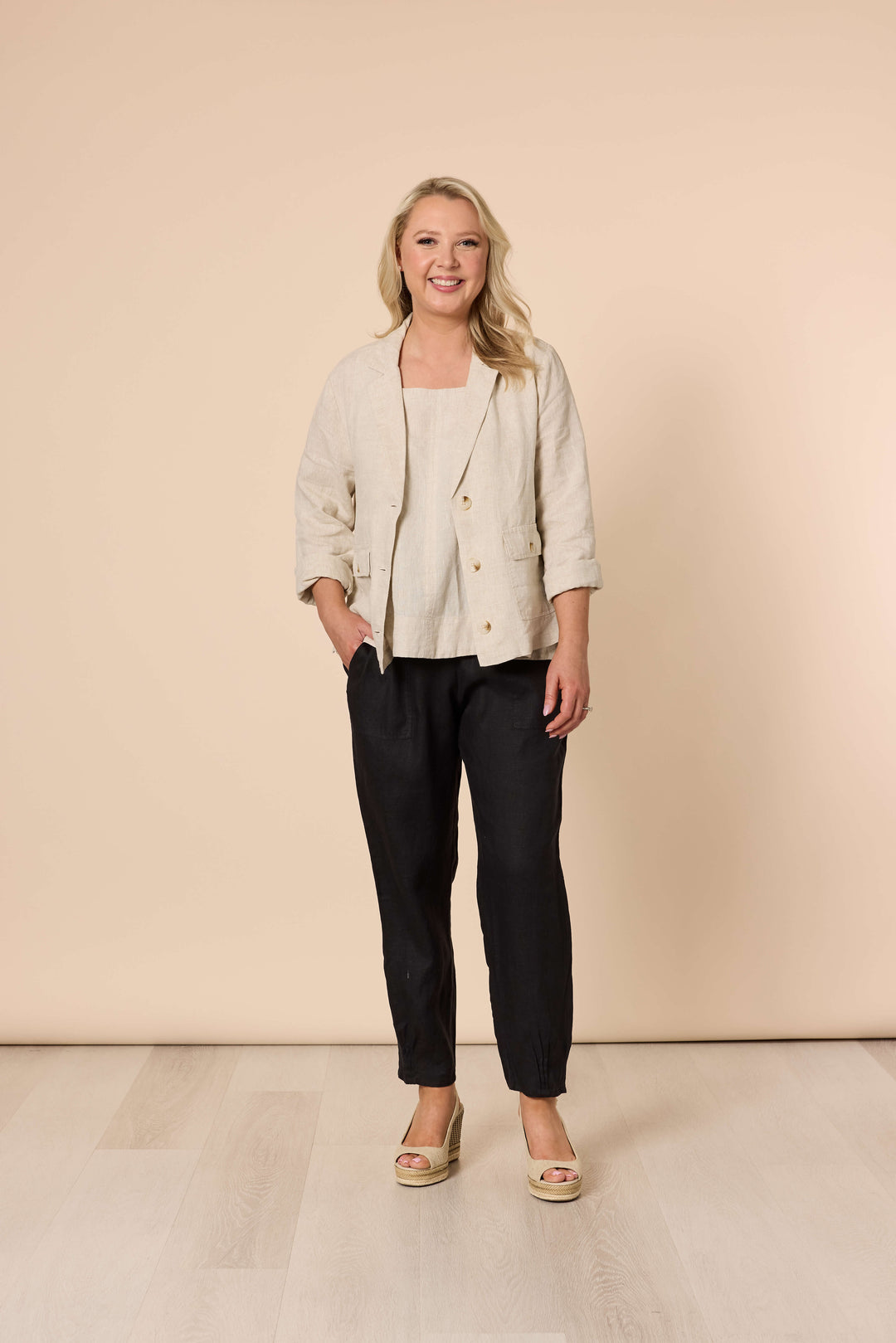 Gordon Smith modeling a light beige linen-blend jacket with black cropped trousers and beige wedge sandals, standing against a solid beige studio backdrop.