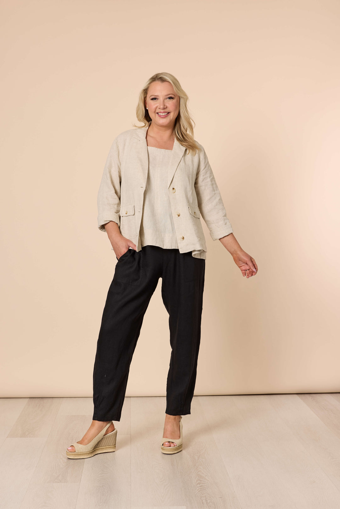 Gordon Smith modeling a light beige linen-blend jacket with black cropped trousers and beige wedge sandals, standing against a solid beige studio backdrop.