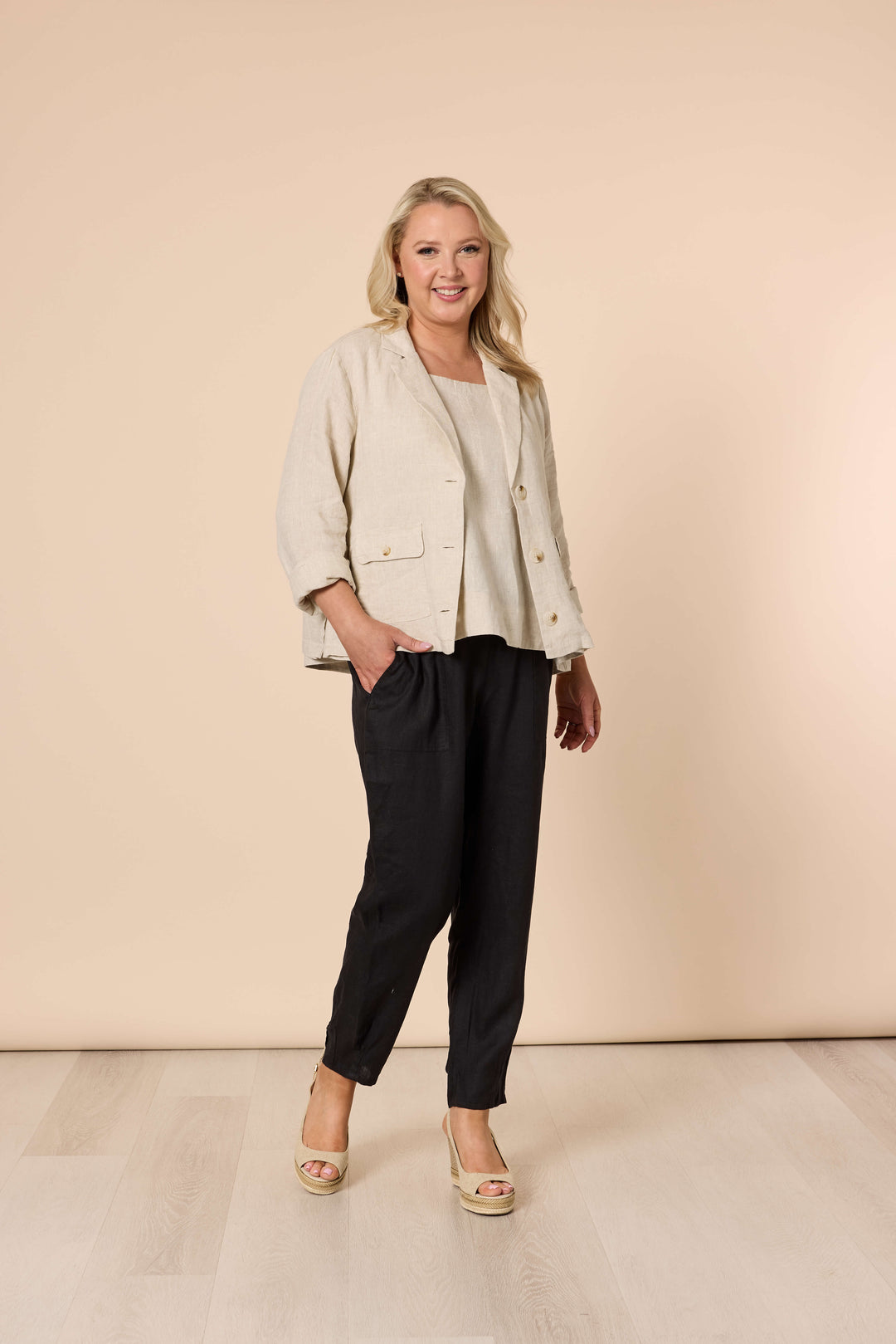 Gordon Smith modeling a light beige linen-blend jacket with black cropped trousers and beige wedge sandals, standing against a solid beige studio backdrop.