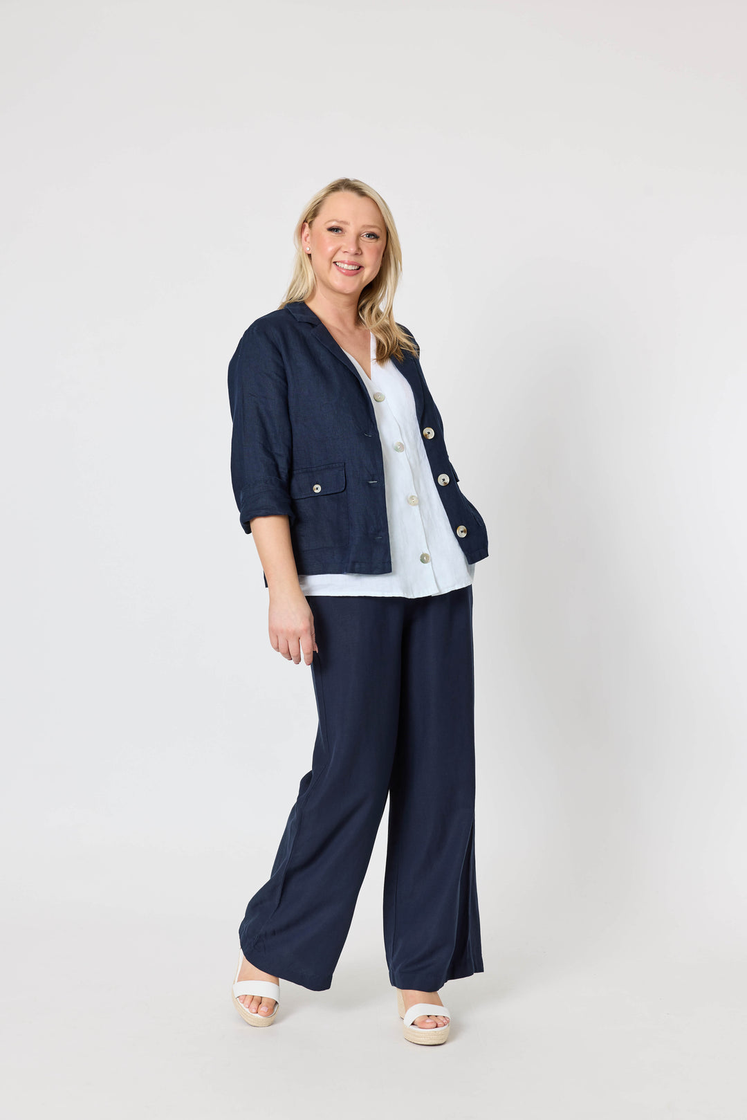 Gordon Smith woman  wearing navy wide-leg pants with a white button-up shirt and white espadrille wedge sandals, standing against a light gray wall and pale wood floor.
