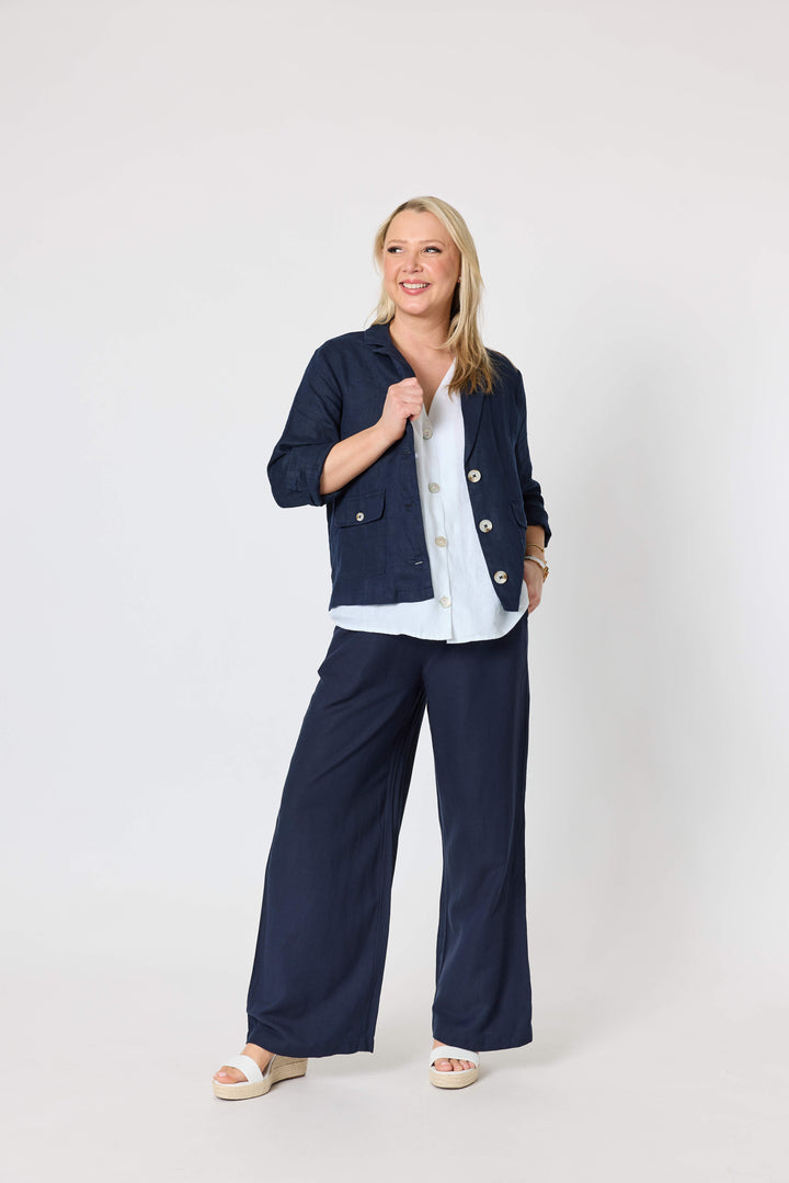 Gordon Smith woman  wearing navy wide-leg pants with a white button-up shirt and white espadrille wedge sandals, standing against a light gray wall and pale wood floor.