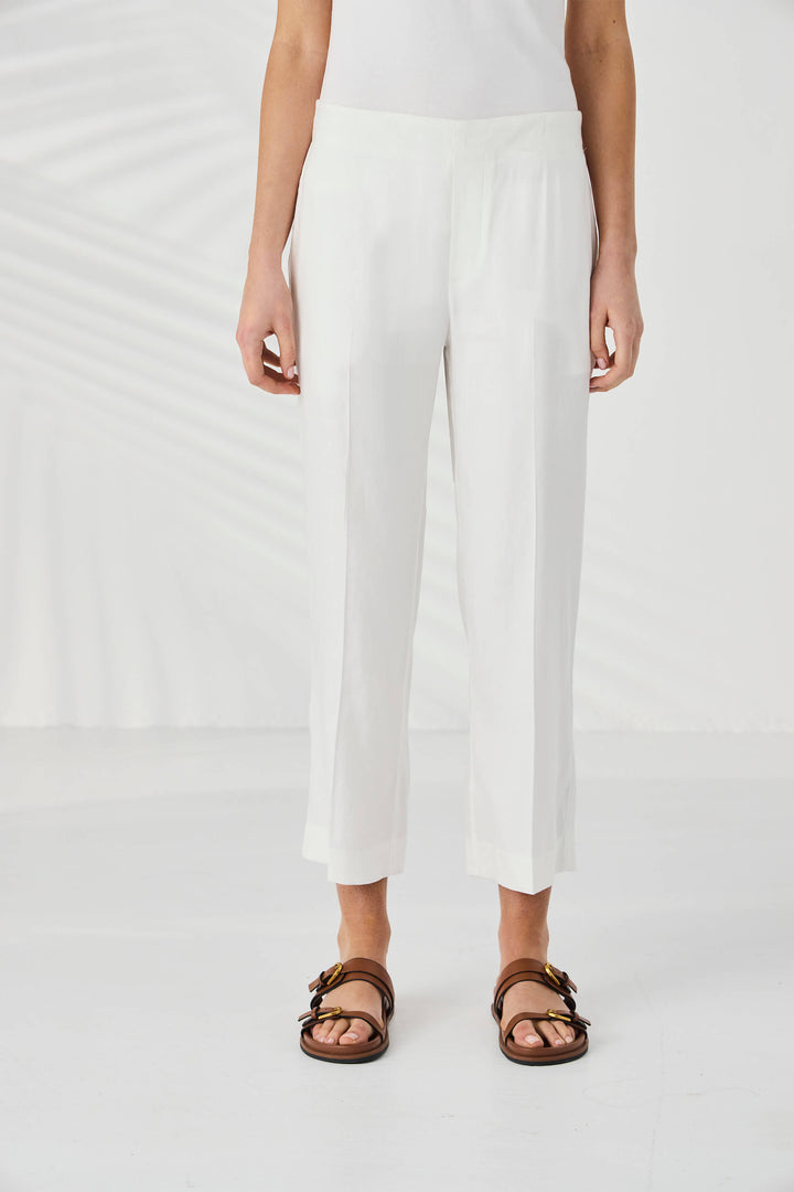 Lania The Label model wearing white cropped trousers with a side slit, styled with a white sleeveless top and brown double-strap sandals against a white background with soft leaf shadows.