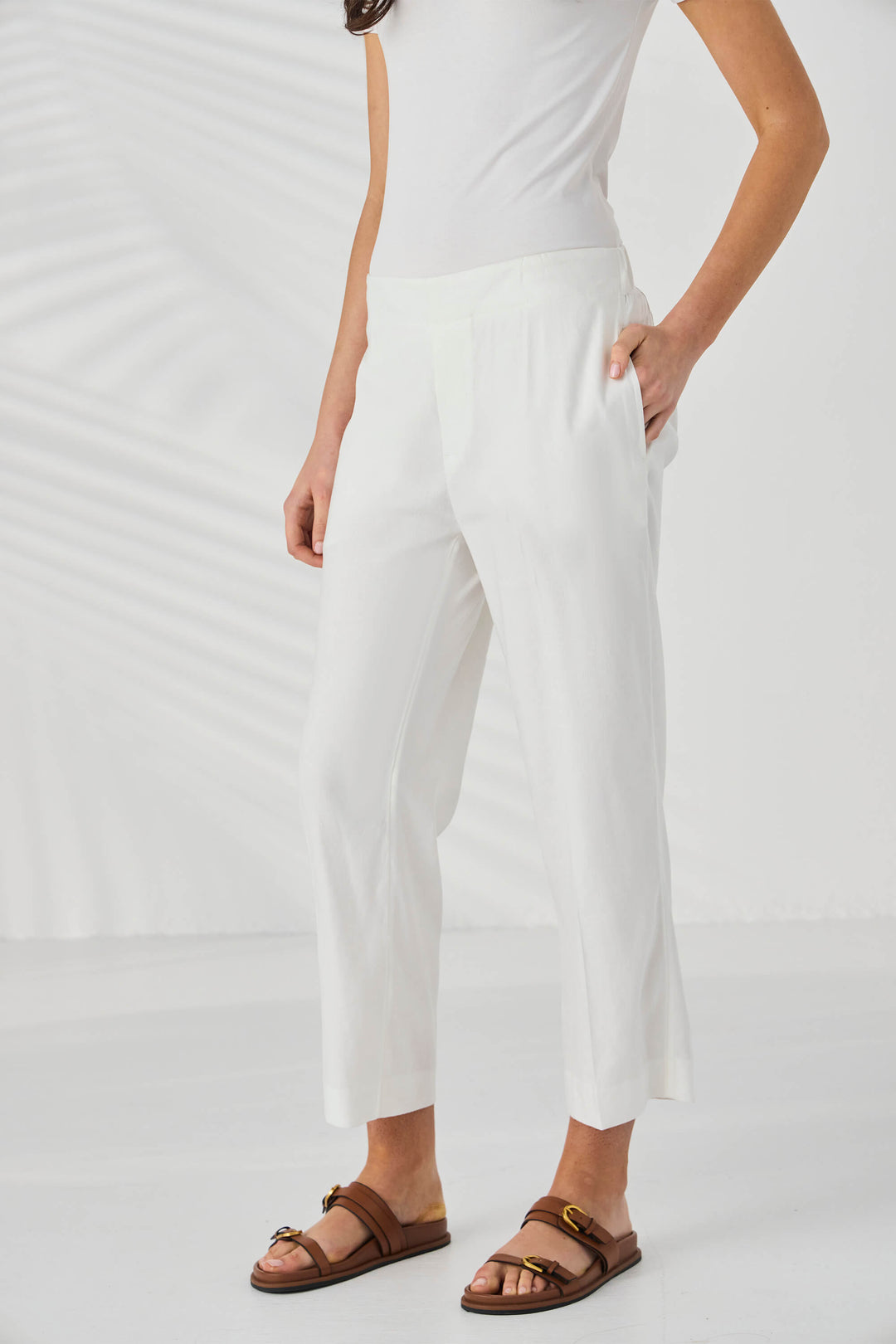 Lania The Label model wearing white cropped trousers with a side slit, styled with a white sleeveless top and brown double-strap sandals against a white background with soft leaf shadows.