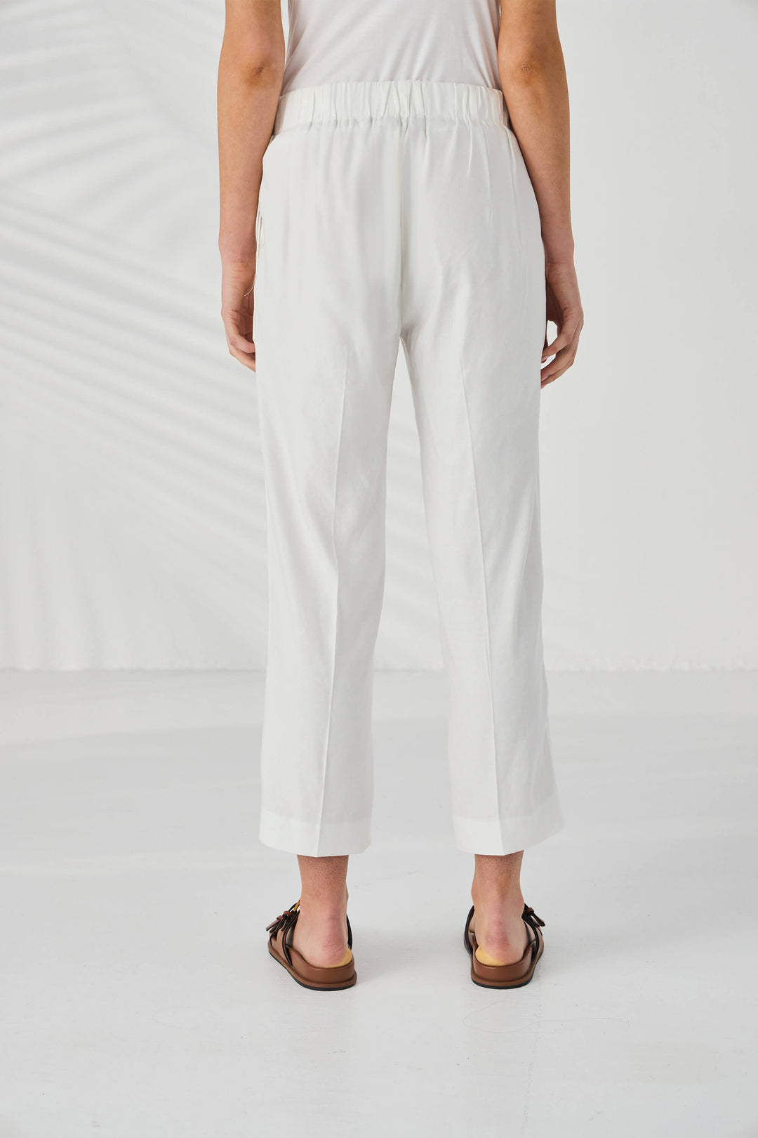 Lania The Label model wearing white cropped trousers with a side slit, styled with a white sleeveless top and brown double-strap sandals against a white background with soft leaf shadows. Back view.