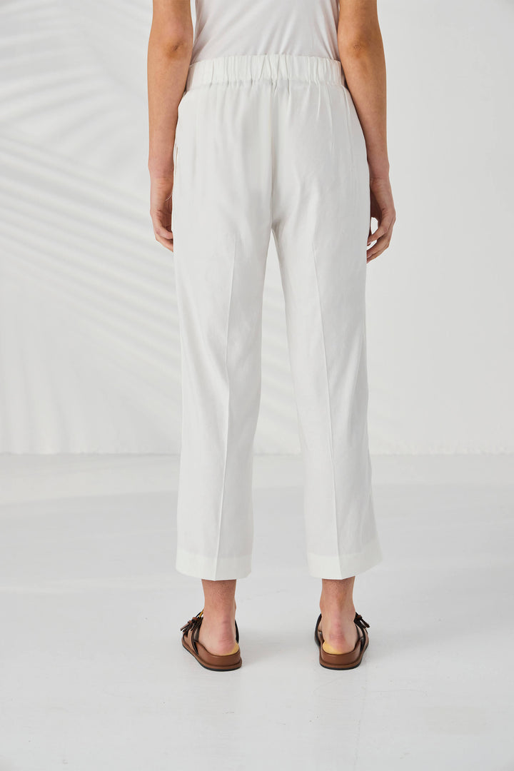 Lania The Label model wearing white cropped trousers with a side slit, styled with a white sleeveless top and brown double-strap sandals against a white background with soft leaf shadows. Back view.