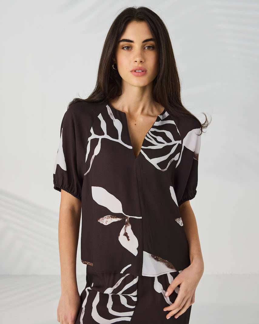 Lania The Label Model modeling a short-sleeve button-front blouse with a dark brown base and large white botanical leaf print, paired with matching dark pants, against a light gray studio backdrop with soft leaf shadows.